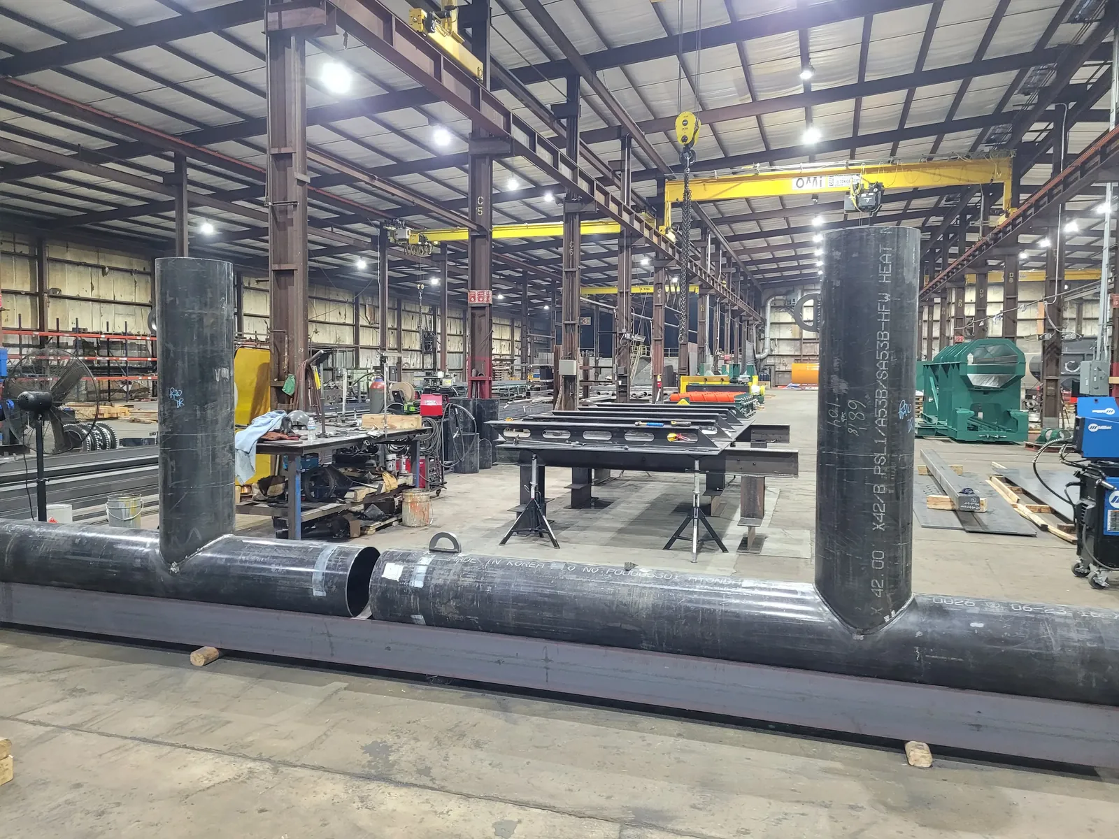 Large-diameter twin-riser pipe weldment laid out in the fabrication bay, bridge cranes overhead